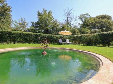 A circular pond with a metal sculpture in the center and two loungers with an umbrella in the background at Utopia in Metfield near Harleston