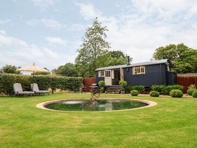 A garden with a circular pond lounge chairs with an umbrella and a dark blue shepherds hut at Utopia in Metfield near Harleston