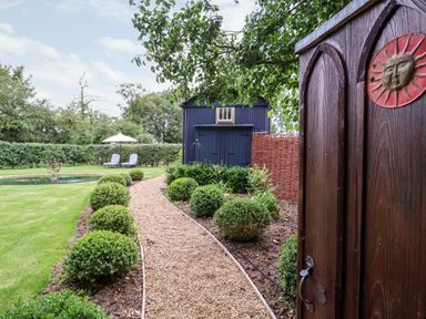 A garden path with trimmed bushes leading to a blue shed and two lounge chairs with an umbrella at Utopia in Metfield near Harleston