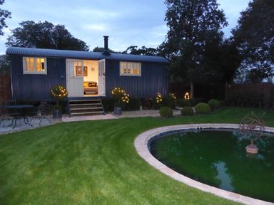 A garden with a grass lawn a circular pond and a small building with open doors and lit windows at Utopia in Metfield near Harleston