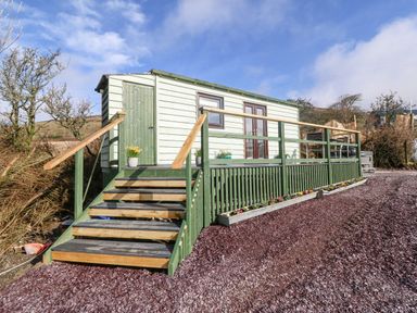 A small wooden cabin with stairs and a railing on a gravel surface surrounded by shrubs and hills at Nyth Y Bioden (Magpie's Nest) in Llanaelhaearn near Trefor