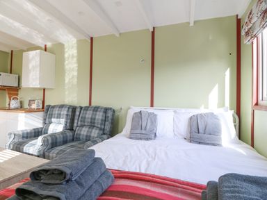 A bedroom with a double bed with gray robes on it a pair of plaid armchairs and a kitchenette with a microwave at Nyth Y Bioden Magpies Nest Llanaelhaearn near Trefor
