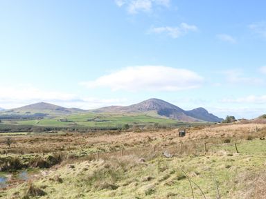 An open field with grass and a wire fence with hills and a partly cloudy sky in the background at Nyth Y Bioden Magpies Nest Llanaelhaearn near Trefor