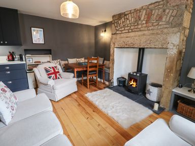 A living room with a wood burning stove a wooden dining table with chairs and light-colored armchairs at Grebe Cottage in Alnmouth