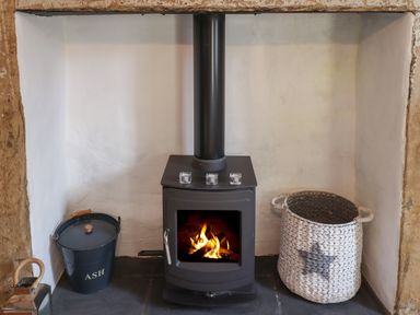 A black wood-burning stove with fire inside placed in a white plastered fireplace with a black ash bucket and a white woven basket at Grebe Cottage in Alnmouth
