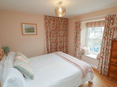 A bedroom with a double bed striped pillows floral curtains a window seat and a wooden chest of drawers at Grebe Cottage in Alnmouth
