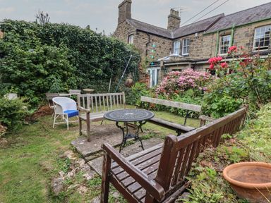A garden with wooden benches a round metal table flowering plants and stone houses at Grebe Cottage in Alnmouth