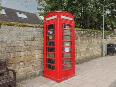 A red telephone booth next to a stone wall with a bench and a litter bin on a paved sidewalk