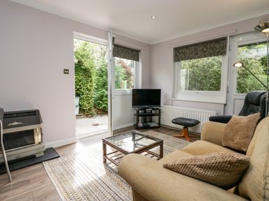 A living room with a beige sofa and a glass coffee table near a TV and open door at The Lodge in Ambleside