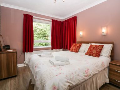 A bedroom with a double bed with white floral bedding red pillows folded towels red curtains wooden bedside table and dresser at The Lodge in Ambleside
