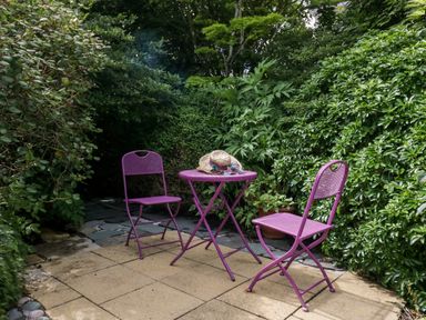 A patio with a purple metal table and two matching chairs with hats on the table surrounded by green bushes at The Lodge in Ambleside