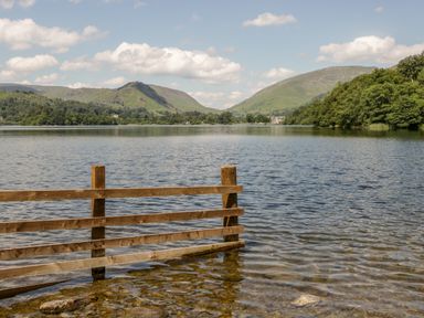 A lake with clear water and a wooden fence in the foreground surrounded by hills and trees at The Lodge in Ambleside