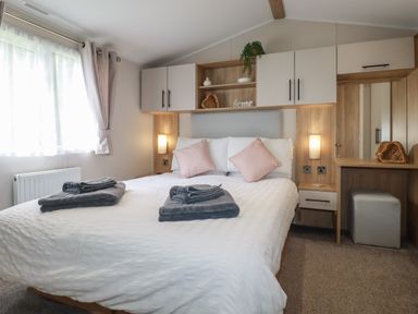 A bedroom with a bed white bedding pink pillows gray towels bedside tables overhead cabinets a dressing table and a window at Pams Place in Crantock