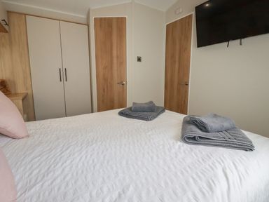 A bedroom with a bed white bedding folded gray towels two wooden doors a wall mounted television and a wardrobe at Pams Place in Crantock