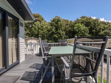 An outdoor deck with a glass table and six chairs overlooking trees and mobile homes at Pams Place in Crantock