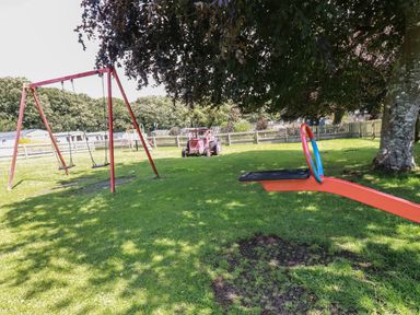 A playground with a swing set a seesaw a toy tractor and a fence with trees in the background at Pams Place in Crantock