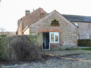 An exterior view of a stone building with a white door and window beside a stone wall covered with dried vines at Luham Cottage Edenhall near Langwathby