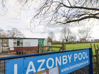 An outdoor view of a pool facility with a small building playground and open field behind a blue gate at Luham Cottage in Edenhall near Langwathby