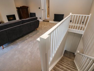 A carpeted upper landing with a dark sofa a wooden cupboard a wall-mounted heater and white stair railings at Luham Cottage in Edenhall near Langwathby