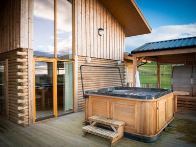 An outdoor wooden hot tub on a deck next to a cabin with large windows at Aurae in Cawdor near Inverness