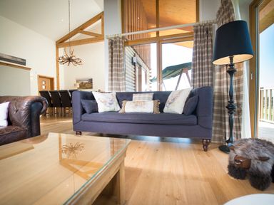 A living room with a blue sofa, a brown leather chair, a wooden coffee table, a black floor lamp, and large windows with plaid curtains at Aurae Cawdor near Inverness