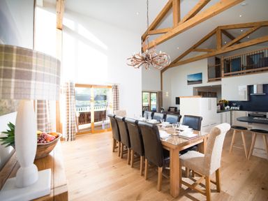 A dining area with a long wooden table set for eight and visible adjoining kitchen with stools at Aurae Cawdor near Inverness