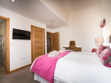 A bedroom with a bed with pink runner and cushions a wooden dressing table a wall mounted tv and wooden doors at Aurae in Cawdor near Inverness