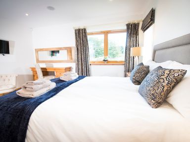 A bedroom with a large bed with folded towels on a blue throw and patterned pillows a desk with a chair a window with curtains a floor lamp and a wall-mounted television at Aurae in Cawdor near Inverness