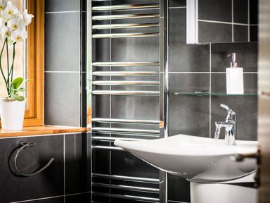 A bathroom with a white sink and faucet a wall-mounted soap dispenser a chrome towel rack a white orchid on a wooden window sill and dark tiled walls at Aurae in Cawdor near Inverness