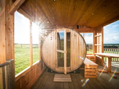 A wooden sauna barrel with an open door inside a wooden shelter with table and benches at Aurae Cawdor near Inverness