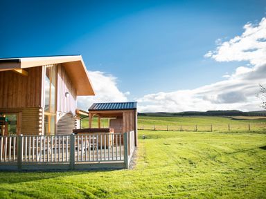 A wooden house with a fenced patio and green fields under a blue sky at Aurae in Cawdor near Inverness