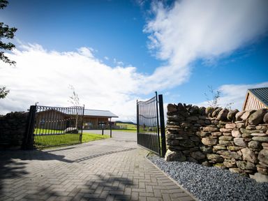 An open metal gate with a stone wall along a paved driveway leading to wooden buildings at Aurae in Cawdor near Inverness