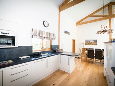A kitchen with white cabinets black countertops a sink and a striped window blind next to a dining area with a wooden table and black chairs at Atlas in Cawdor near Inverness