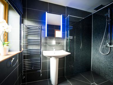 A bathroom with a white pedestal sink a mirror with lights a chrome towel rack and a walk in shower with black tiled walls and a glass partition at Atlas in Cawdor near Inverness
