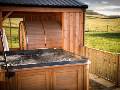 An outdoor hot tub on a wooden deck with a structure and green field in the background at Atlas in Cawdor near Inverness