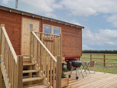 A wooden hut on wheels with stairs leading to the door and a deck with a barbecue and table with two chairs at Shepherds Hut in Garthorpe near Scunthorpe