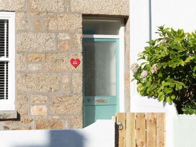 A stone wall with a green door and a red heart house number plate 11 with a wooden gate and plants at 11 Belgravia Street in Penzance