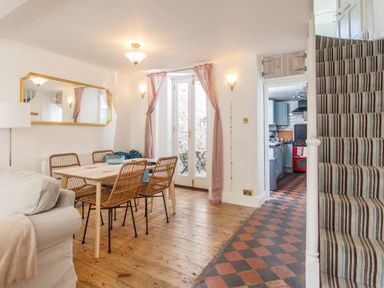 A dining area with a wooden table and wicker chairs next to glass doors with curtains and a view into a kitchen and striped carpeted stairs at 11 Belgravia Street in Penzance