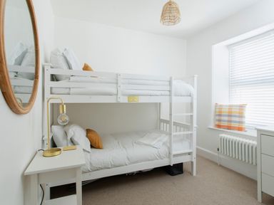 A bedroom with white bunk beds a small white bedside table with a gold lamp a round mirror on the wall a window with blinds and a plaid cushion on the sill at 11 Belgravia Street in Penzance