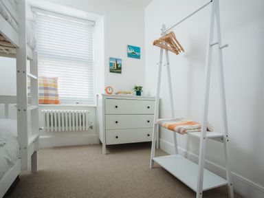 A small bedroom with a white bunk bed a window with blinds a white dresser with books and a clock a white clothing rack with hangers and a folded blanket at 11 Belgravia Street in Penzance