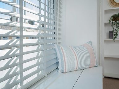 A window with white blinds and a striped pillow on the windowsill near a shelf with plants at 11 Belgravia Street in Penzance