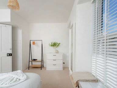 A bedroom with a bed, a chest of drawers with a plant on top, a clothing rack with hangers, and a window with blinds and a folded blanket on the sill at 11 Belgravia Street in Penzance