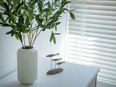 A white vase with green leafy branches and a wooden fish decoration on a white surface near white window blinds at 11 Belgravia Street in Penzance
