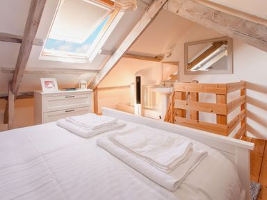 A bedroom with a skylight ceiling window a wooden bed with white linens a dresser with books and a lantern and a wooden railing at 11 Belgravia Street in Penzance