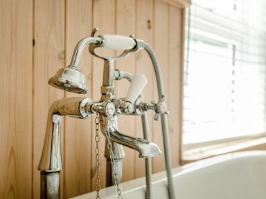 A close up of a vintage bathtub faucet and shower head with a wooden wall and window blinds in the background at 11 Belgravia Street in Penzance