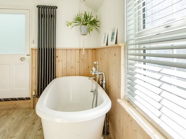 A bathroom with a white freestanding bathtub wooden panel walls a tall black radiator a hanging plant and window blinds at 11 Belgravia Street in Penzance