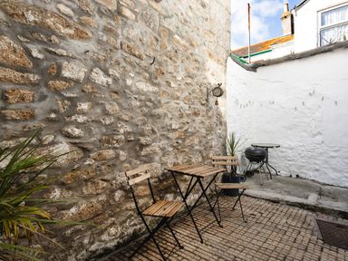 A small outdoor patio with a wooden table and two chairs a barbecue grill plants and stone and white walls at 11 Belgravia Street in Penzance