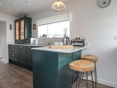 A kitchen with green cabinets a microwave a toaster two wooden stools and a window with blinds at Bwthyn Y Swnt in Moelfre