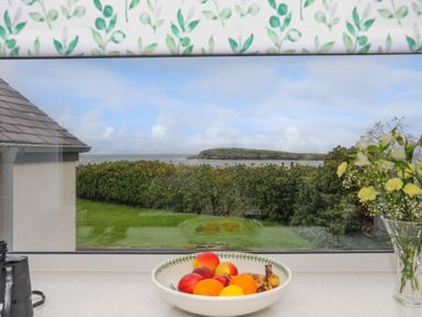 A kitchen countertop with a bowl of fruit and a vase of flowers by a window showing a garden and sea view at Bwthyn Y Swnt in Moelfre