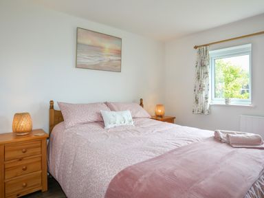 A bedroom with a bed with pink bedding two wooden bedside tables with lamps and a window with floral curtains at Bwthyn Y Swnt in Moelfre